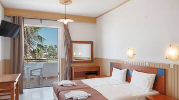 A hotel room with a bed, bedside tables, a desk, a TV, a balcony with chairs, and a view of palm trees.
