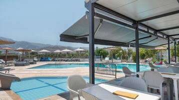 A poolside area with tables and chairs, a covered seating area, and a view of mountains in the background.