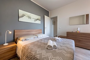 A modern bedroom with a large bed, a bedside lamp, a wooden dresser, and a wall-mounted mirror. - Apartment 'My Town Home' with Balcony, Wi-Fi and Air Conditioning (Torri del Benaco)