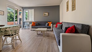 A living room with a sofa, a dining table, and a chair. There is a window with blinds, a picture on the wall, and a door leading to another room. - Apartment 'My Town Home' with Balcony, Wi-Fi and Air Conditioning (Torri del Benaco)
