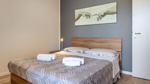 A neatly made bed with a patterned bedspread and two rolled towels, a wooden headboard, a wall-mounted artwork, and a bedside lamp. - Apartment 'My Town Home' with Balcony, Wi-Fi and Air Conditioning (Torri del Benaco)