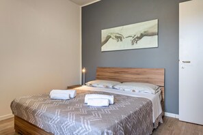 A neatly made bed with a patterned bedspread and two rolled towels, a wooden headboard, a wall-mounted artwork, and a bedside lamp. - Apartment 'My Town Home' with Balcony, Wi-Fi and Air Conditioning (Torri del Benaco)