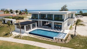 A modern two-story house with a swimming pool, large windows, and a view of the ocean. - Coral Cove Villa - New Large 7 Bedroom Modern Luxury Villa + Concierge (George Town)