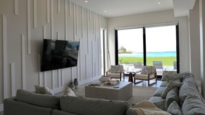 A modern living room with a large flat-screen TV, a grey sofa, a coffee table, and a view of the ocean through large windows. - Coral Cove Villa - New Large 7 Bedroom Modern Luxury Villa + Concierge (George Town)