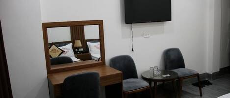 A hotel room with a wooden desk, two grey chairs, a small round table with a glass, a flat-screen TV mounted on the wall, and a bed with a white mattress.