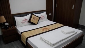 Una cama bien hecha con una colcha blanca y una manta color beige, dos almohadas con el logotipo del hotel, y una mesita de noche de madera con una lámpara.
