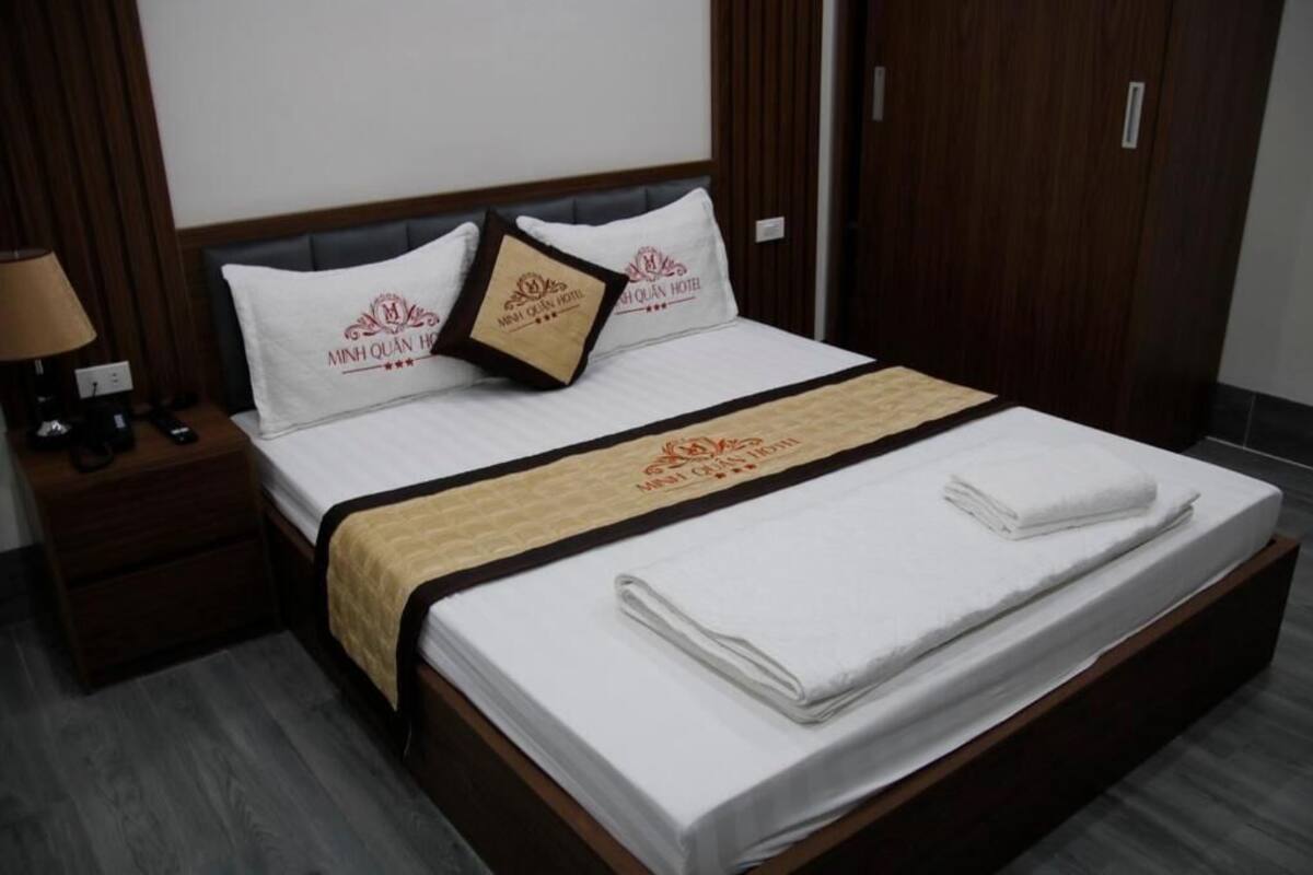 Una cama bien hecha con una colcha blanca y una manta color beige, dos almohadas con el logotipo del hotel, y una mesita de noche de madera con una lámpara.