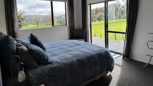 A bedroom with a bed, a chair, a desk, and a view of a green landscape through large windows. - Nibble Nook Farm Cottage (Tapapa)