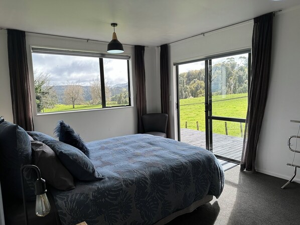A bedroom with a bed, a chair, a desk, and a view of a green landscape through large windows.