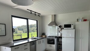 A modern kitchen with white cabinetry, a large window with a view of greenery, a gas stove, a microwave, and a refrigerator. - Nibble Nook Farm Cottage (Tapapa)