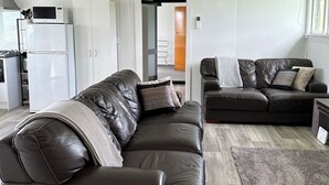A modern living room with two leather sofas, a kitchen area with white cabinets, and a grey shag rug. - Nibble Nook Farm Cottage (Tapapa)