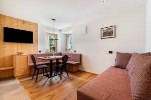Una habitación con una pared de madera y un televisor, un comedor con mesa y sillas, un sofá, y una ventana con cortinas.