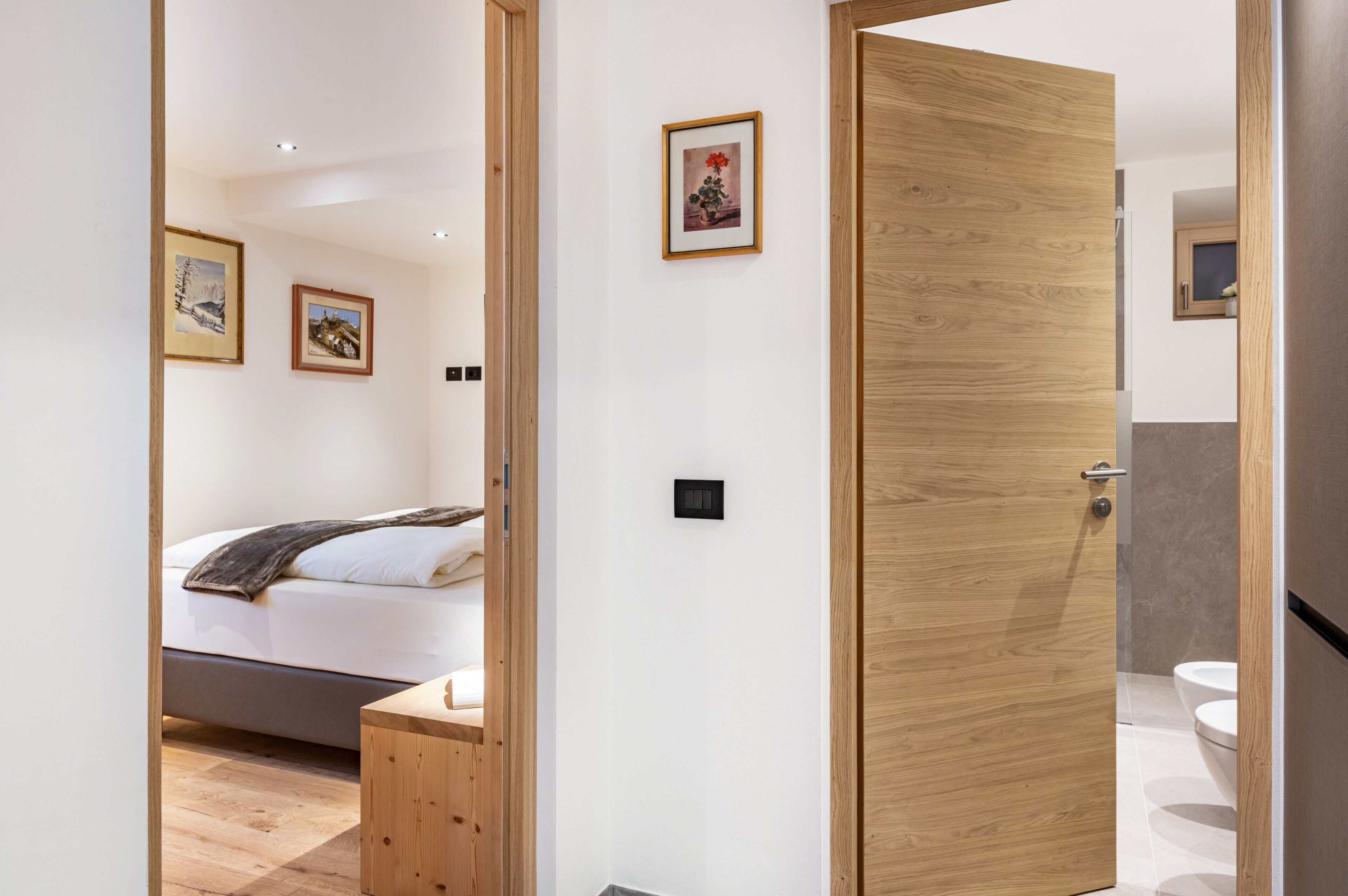 Une chambre d’hôtel moderne avec une porte en bois, un lit avec un matelas blanc, une petite table de chevet en bois et un tableau encadré au mur.