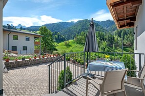 A balcony with a table and chairs, a view of a mountainous landscape, and a building with a sloped roof.