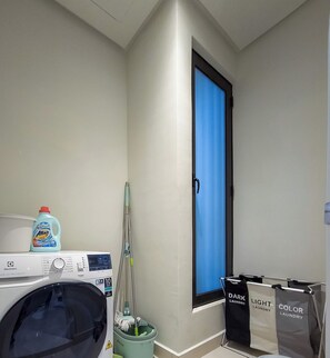 A laundry area with a washing machine, a bucket, and laundry bags labeled 'DARK', 'LIGHT', and 'COLOR'.
