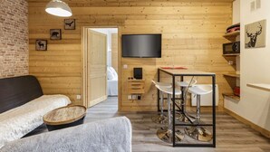 Une pièce chaleureuse avec un plafond en bois, un téléviseur fixé au mur, un coin repas avec une table et des chaises, et un canapé agrémenté d’un plaid moelleux.