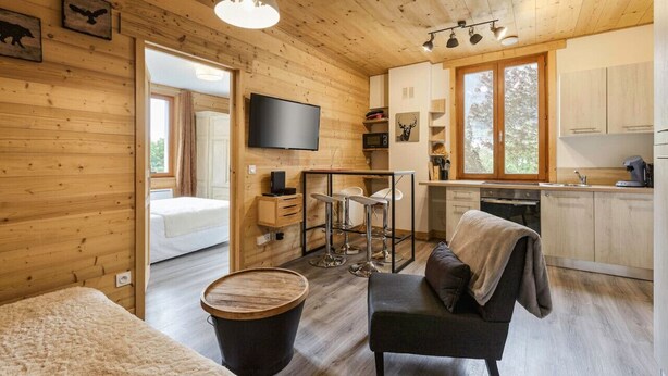 L’intérieur chaleureux d’un chalet, avec un revêtement mural en bois, un téléviseur fixé au mur, un coin repas avec des tabourets et une cuisine équipée d’armoires.