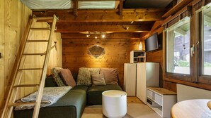 A cozy cabin interior with a wooden staircase, a green sofa, a white ottoman, and a television on a wooden cabinet.
