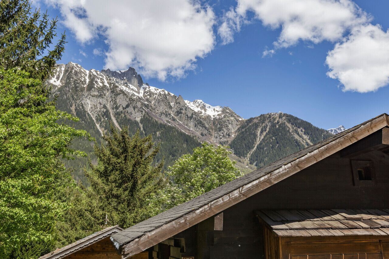 Une cabane en bois, dotée d’un toit en pente, entourée d’une végétation luxuriante et avec des montagnes en arrière-plan.