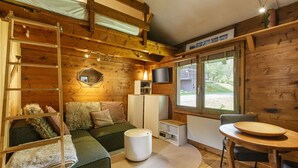 A cozy cabin interior with a loft bed, a sofa, a round table, and a TV mounted on the wall.