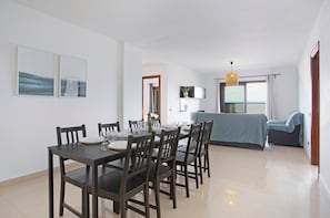 Uma sala de jantar com uma mesa posta para quatro pessoas, um sofá, uma televisão e uma janela com cortinas.