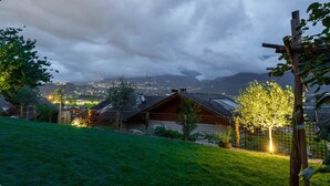 Une maison de campagne avec un toit en pente, entourée d’une cour clôturée et d’arbres.