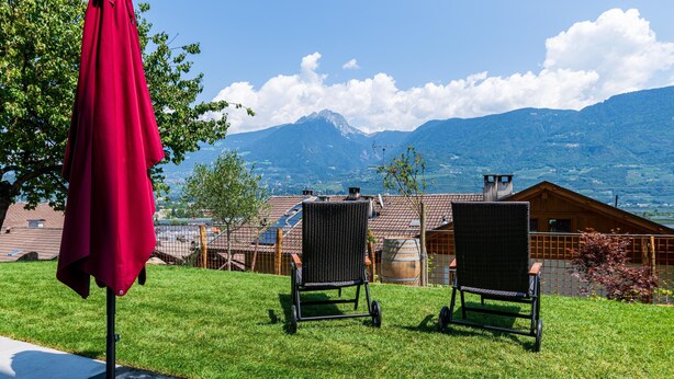 Un espace extérieur verdoyant avec deux chaises et un parasol rouge, offrant une vue sur une montagne et un bâtiment en bois.