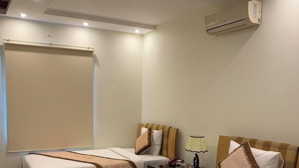 A hotel room with two beds, a ceiling fan, an air conditioning unit, a bedside table with a lamp, and a window with a blind. - Love Hotel Hoang Mai HN- by Bay Luxury (Hanoi)