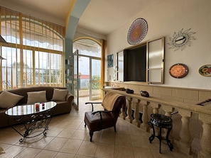 A room with a sofa, a table, a chair, a fireplace, and a view through large windows.