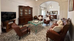 A living room with a television, wooden cabinet, leather chairs, a glass table, and a chandelier.