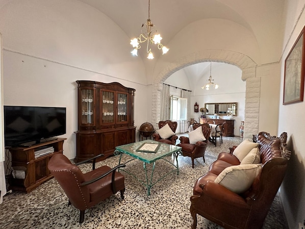A living room with a television, wooden cabinet, leather chairs, a glass table, and a chandelier.