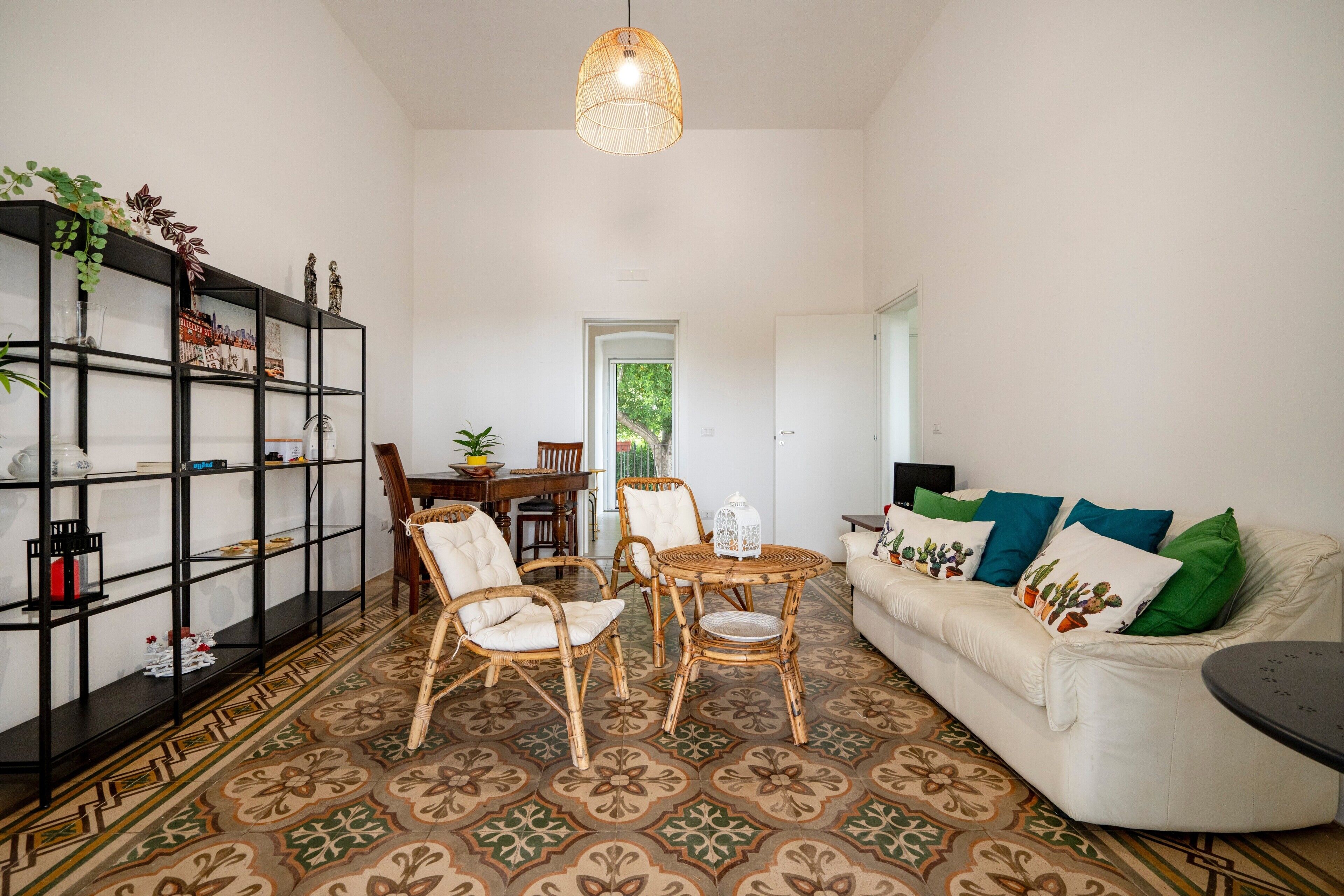 A living room with a white sofa, a dining table, wicker chairs, a patterned floor, a bookshelf, and a hanging light fixture.