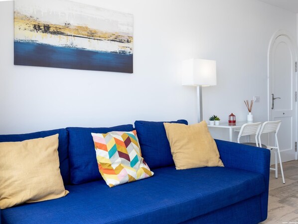 Un sofá azul con dos cojines color beige y un almohadón geométrico de colores, una lámpara blanca, una mesita con un farol rojo y una silla blanca.
