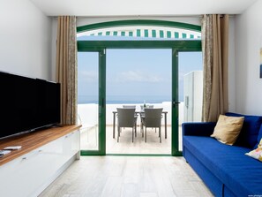 Una habitación moderna con televisión, un sofá azul, una mesa de comedor con sillas y vistas al mar a través de una puerta de cristal.