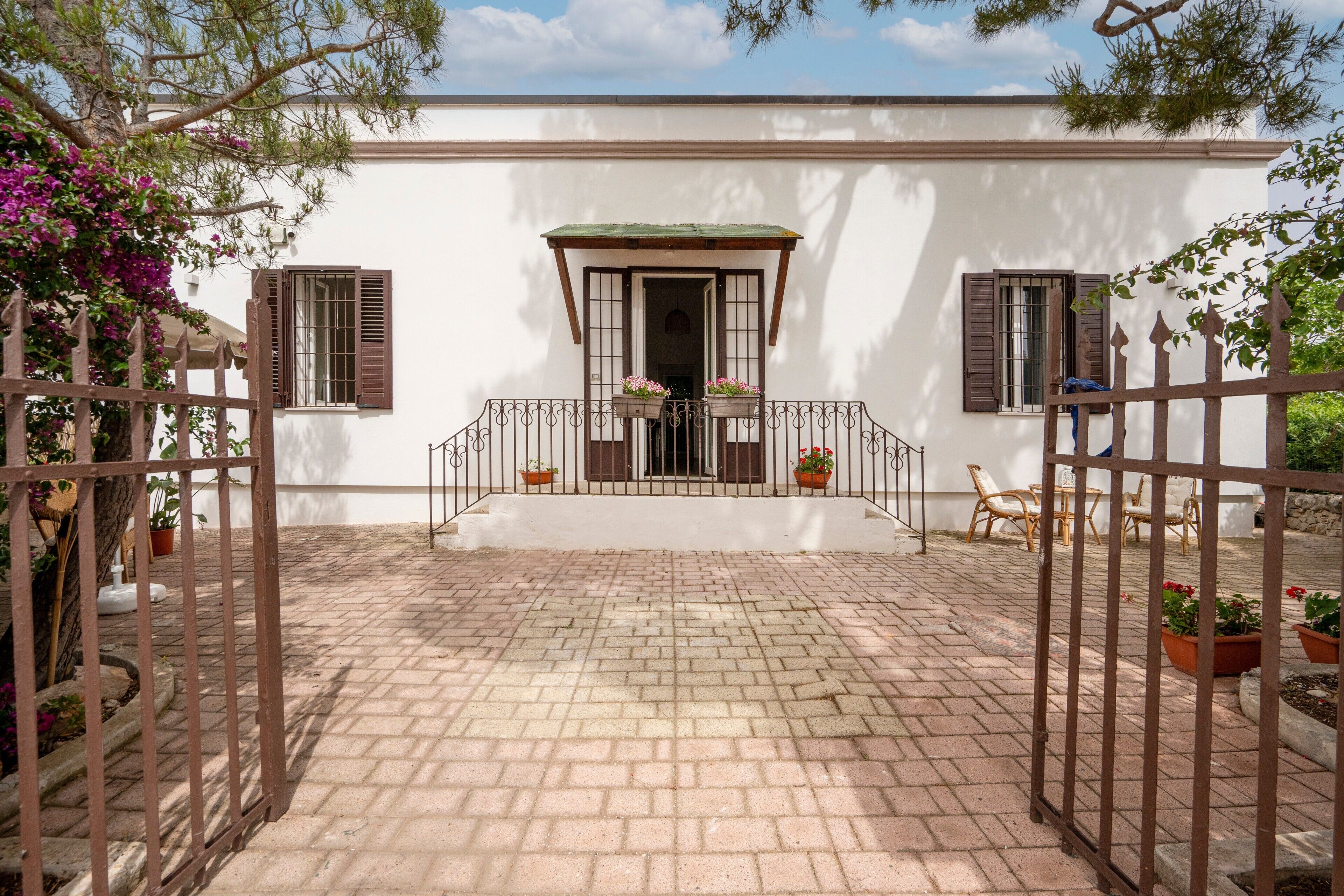 A white building with a brick-paved entrance, a gated porch with potted plants, and a clear sky.