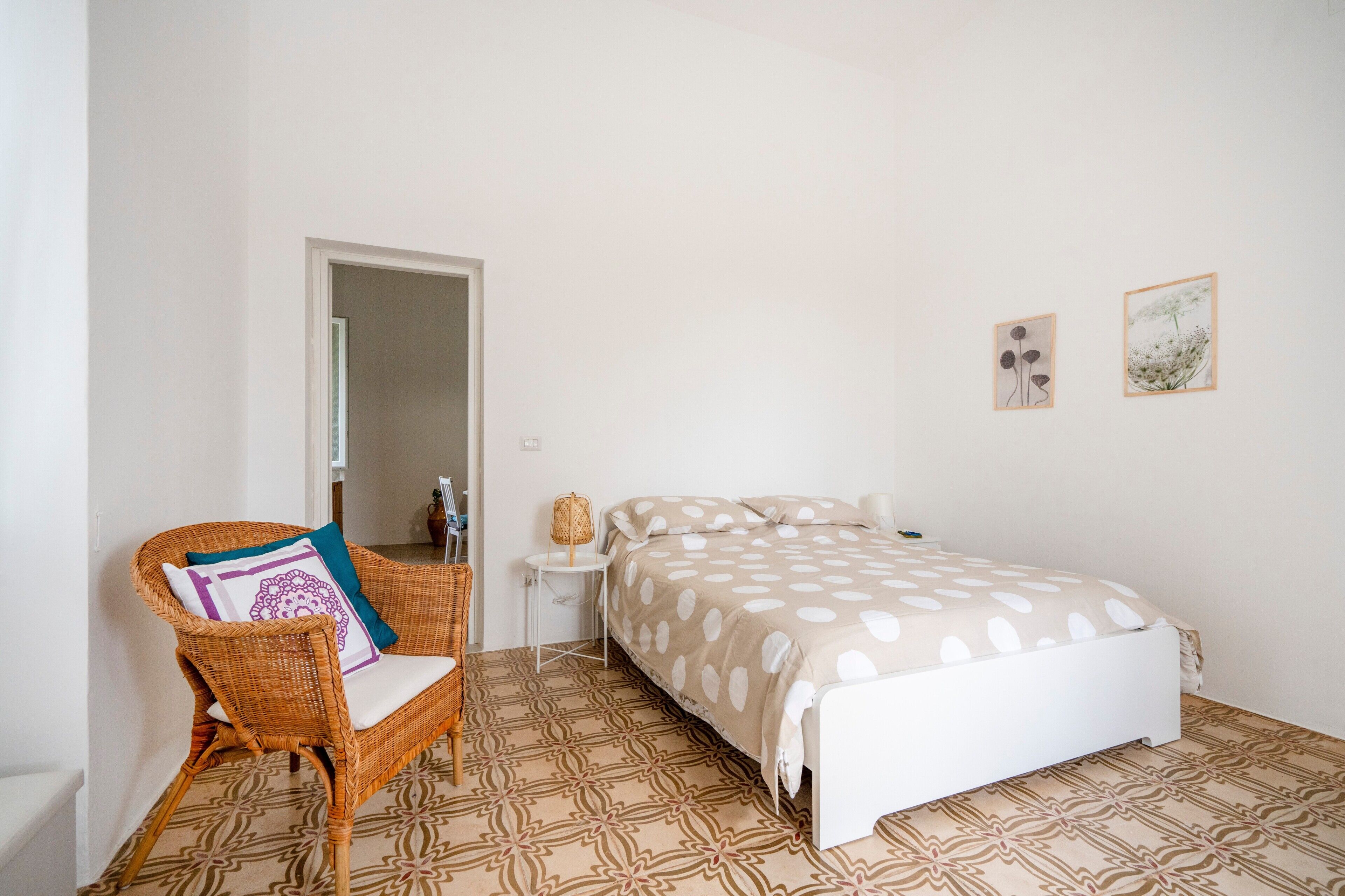 A bedroom with a bed, a wicker chair with a cushion, a small bedside table, and a patterned floor.