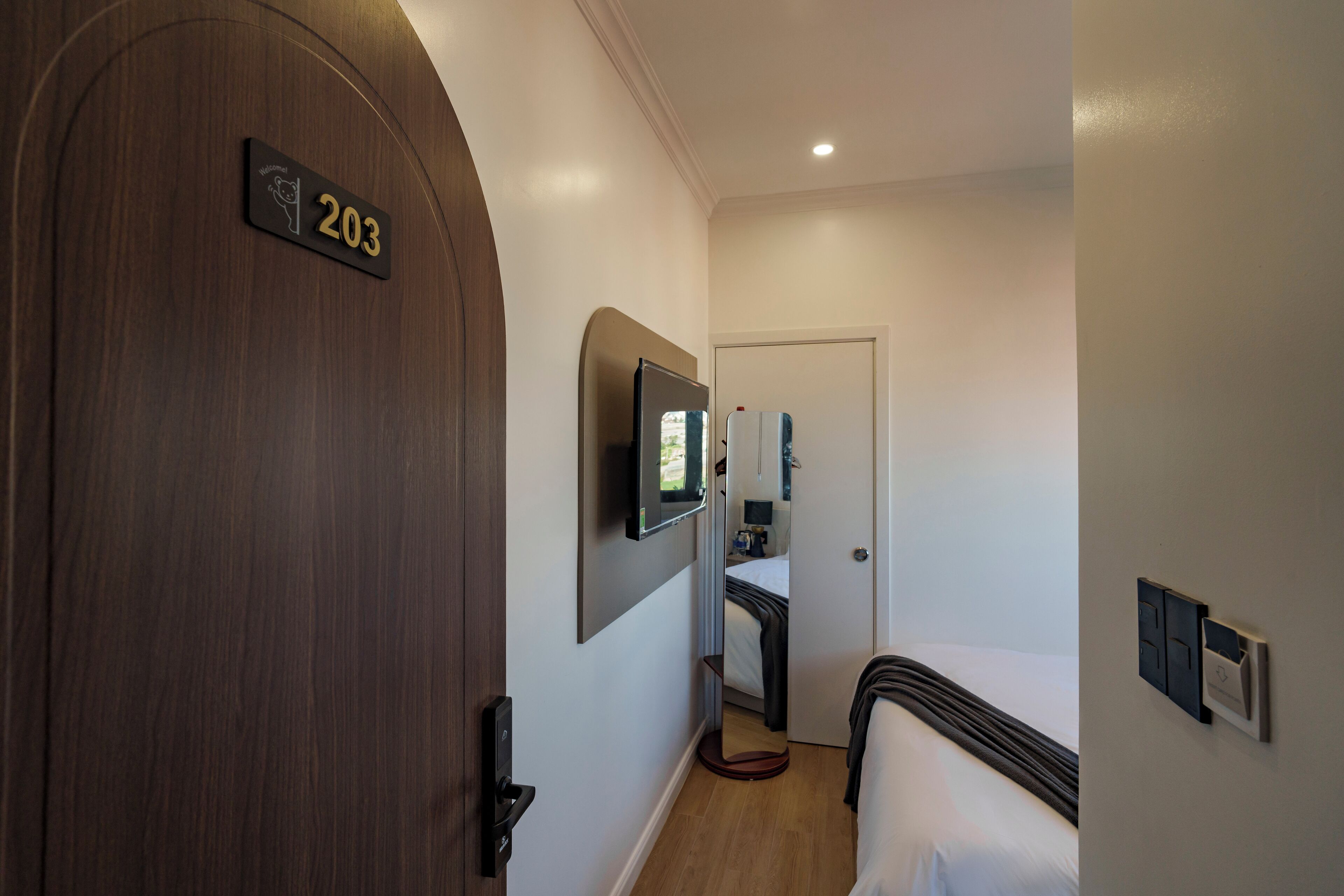 A hotel room with a wooden door labeled "203", a bed, a TV mounted on the wall, and a bedside table with a lamp.
