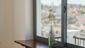 A modern room with a large window, a wooden table, two bar stools, and a vase with plants on the windowsill. - Storij Home & Coffee (Lam Vien - Da Lat)