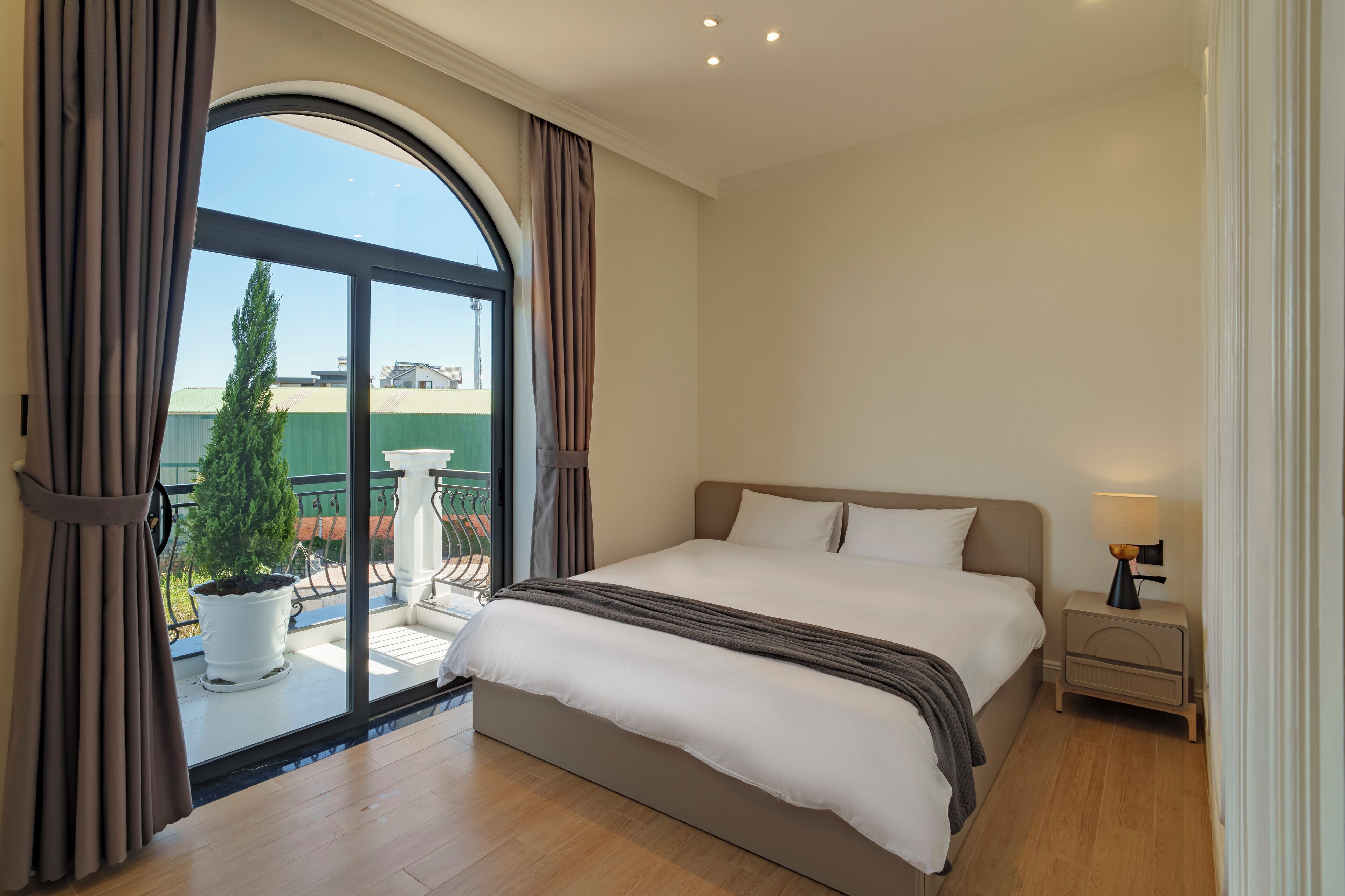 Ein Schlafzimmer mit einem großen Bett, einem Nachttisch mit Lampe, einer Topfpflanze und Blick auf ein Gebäude durch ein großes Fenster.