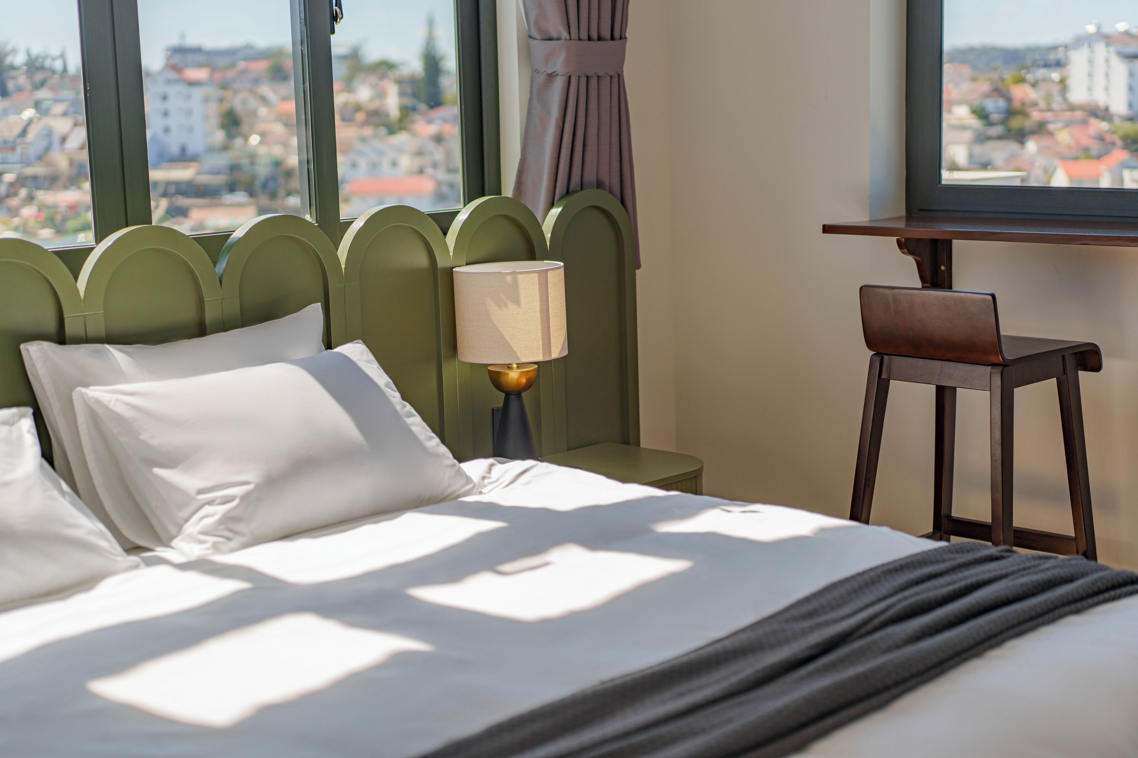 Ein Hotelzimmer mit einem Bett, einem Nachttisch, einer Lampe und Blick auf die Stadt durch große Fenster.