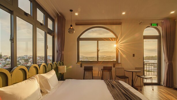 A hotel room with a large bed, a desk with two chairs, a window with a view of the city, and an exit sign. - Storij Home & Coffee (Lam Vien - Da Lat)