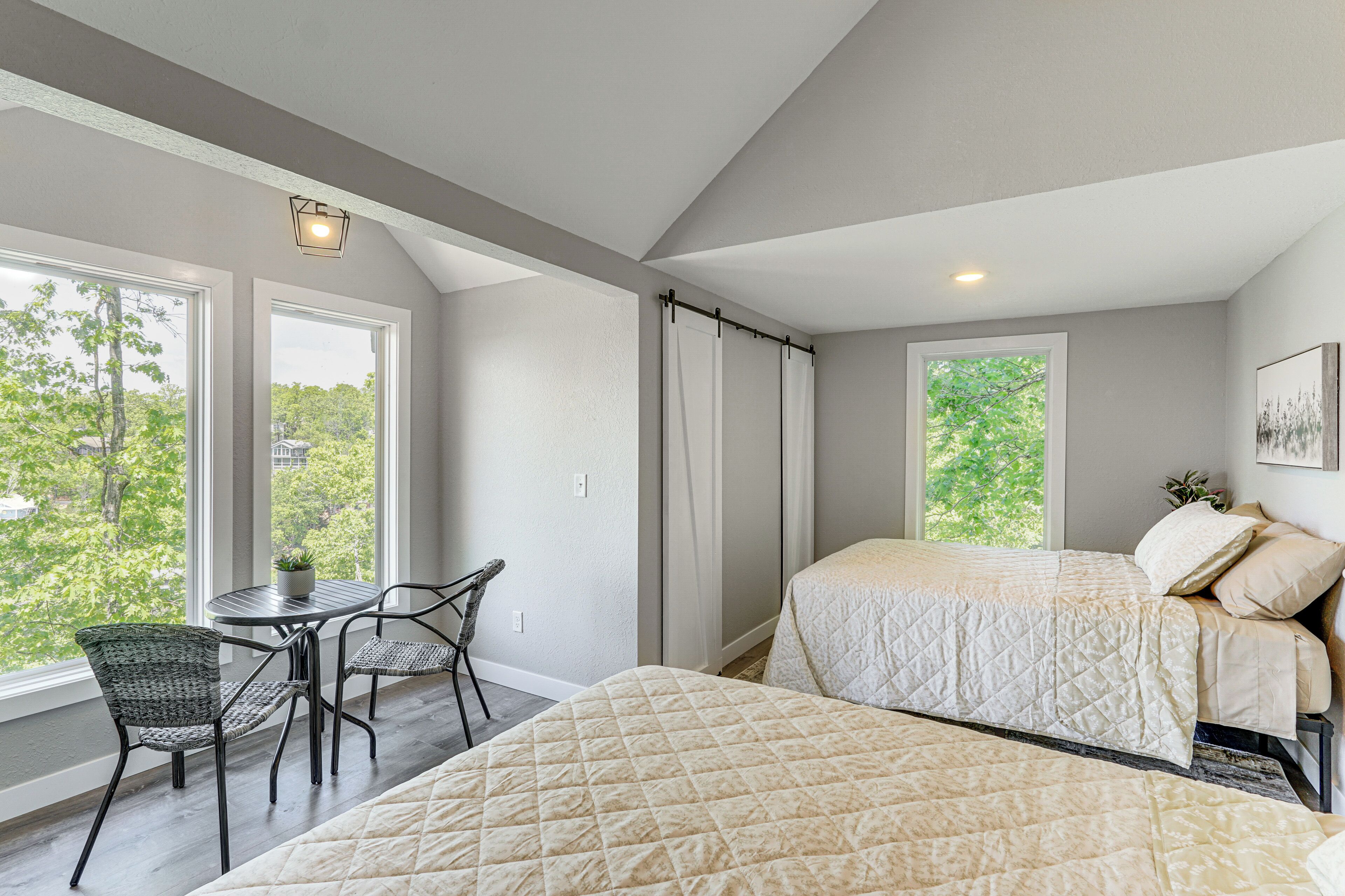 A bedroom with two beds, a dining table with chairs, and large windows offering a view of trees.