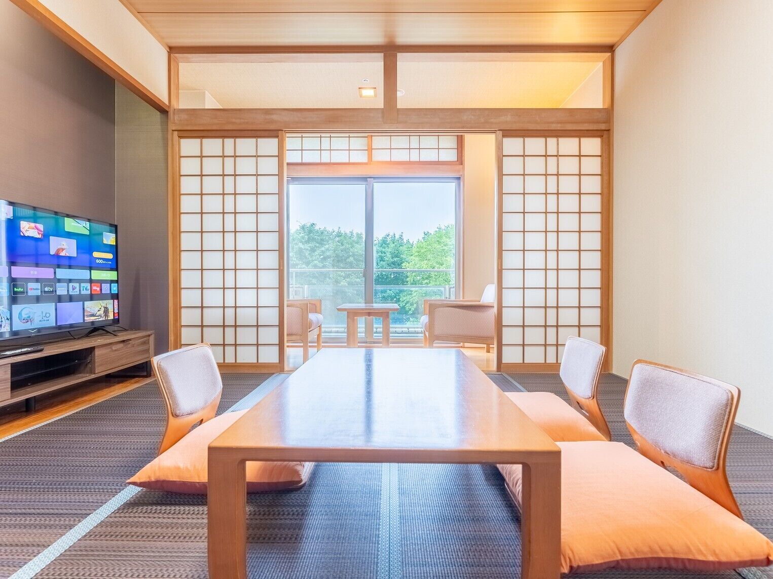Habitación de hotel con un amplio ventanal corredero, un televisor de pantalla plana, una mesa de madera y dos sillones acolchados.