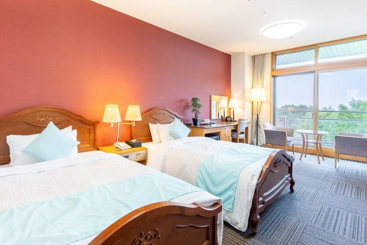 Een hotelkamer met twee bedden, een bureau met stoel, een groot raam en uitzicht op groen.