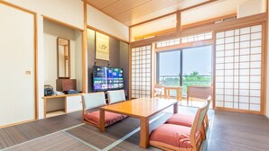 Ein traditioneller japanischer Raum mit Tatami-Boden, einem niedrigen Tisch und Kissen. Ein Fernseher ist an der Wand befestigt, und es gibt eine Schiebetür zu einem Balkon.