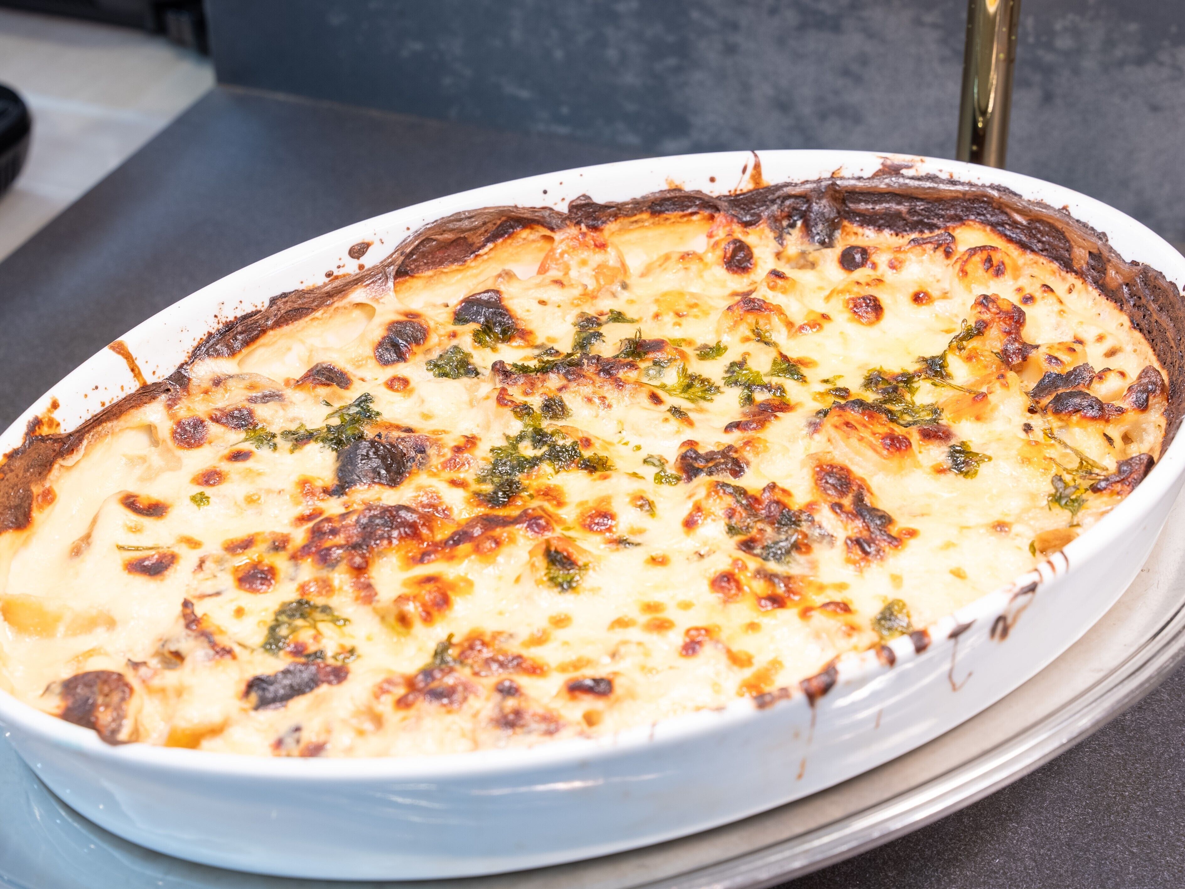 A white oval dish with a golden-brown crust, likely a baked dish with cheese and herbs, on a dark countertop.