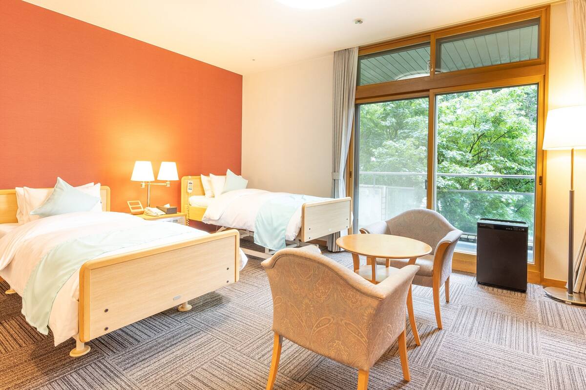 Een hotelkamer met twee bedden, een klein tafeltje, een stoel, een lamp en een groot raam met uitzicht op bomen.
