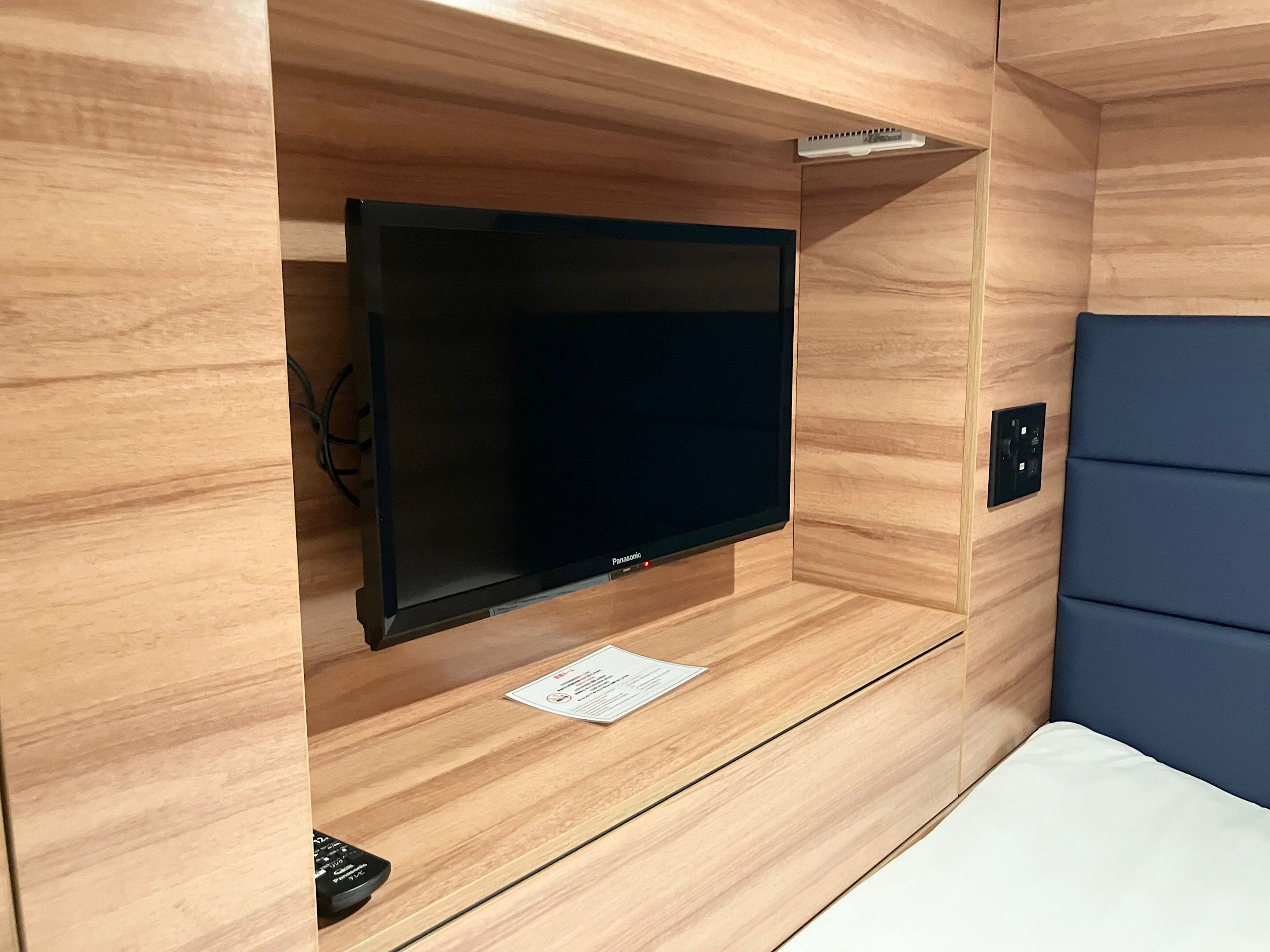 Quarto compacto com televisão de tela plana montada, painel de controle e uma tomada na parede.