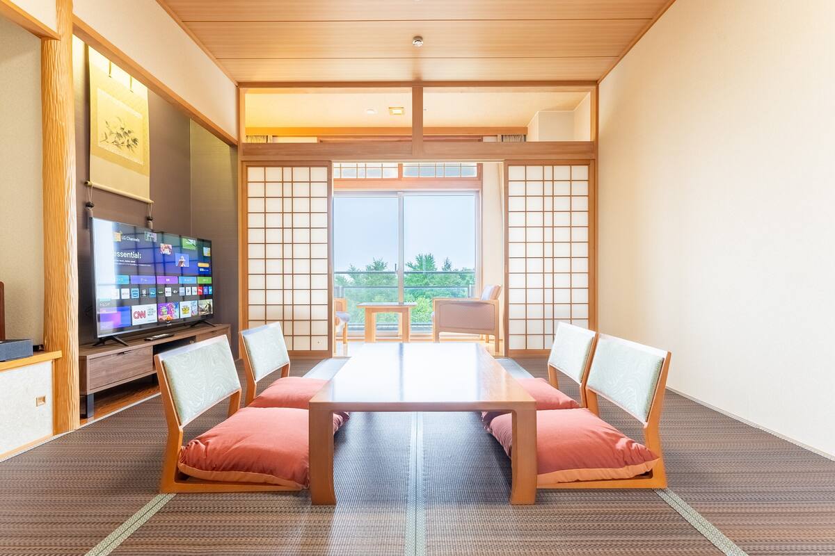 Een traditionele Japanse kamer met tatami vloeren, een lage tafel met kussens, een televisie op een houten standaard, en een schuifdeur naar een balkon.