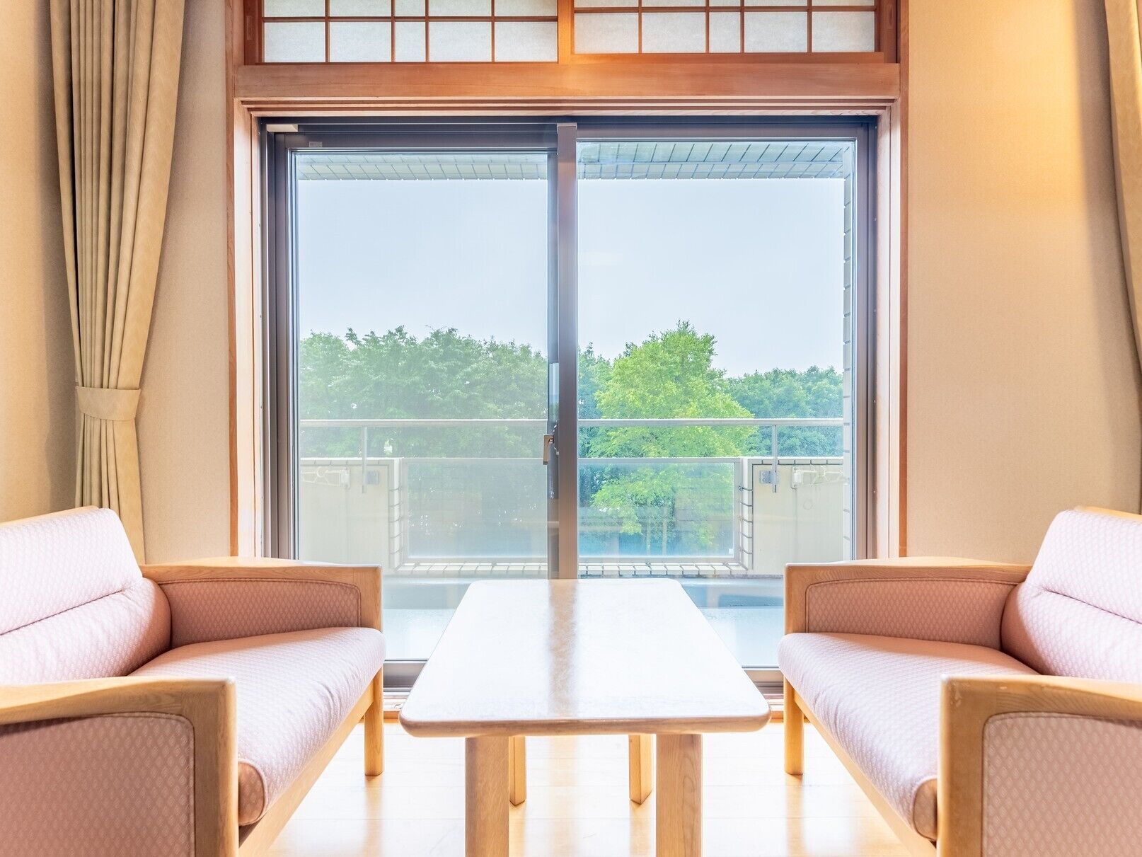 Una habitación con un amplio ventanal corredera, dos sillones acolchados, una mesa de madera y vistas al exterior con vegetación.